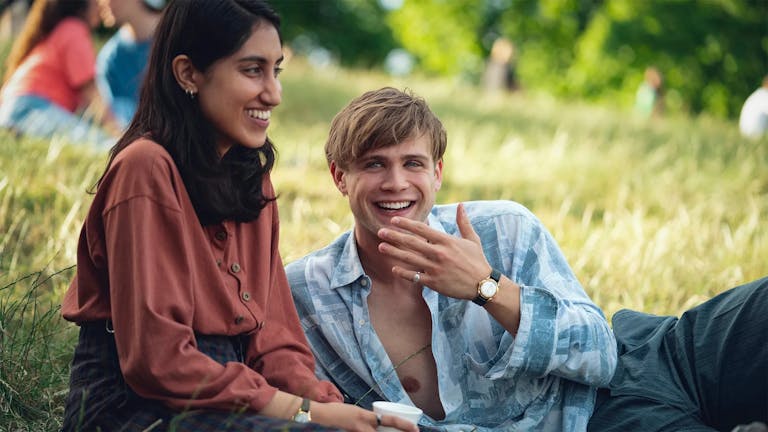 A young brown woman sits on a sunny grassy field next to a young white man lounging beside her, both smiling 
