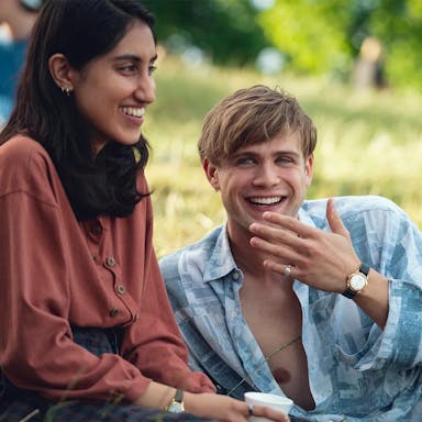 A young brown woman sits on a sunny grassy field next to a young white man lounging beside her, both smiling 