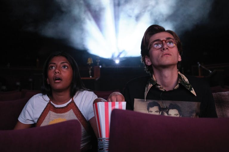 A brown teenage girl sits next to a white teenage boy in a cinema screen eating popcorn looking shocked at the screen