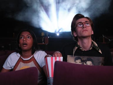A brown teenage girl sits next to a white teenage boy in a cinema screen eating popcorn looking shocked at the screen