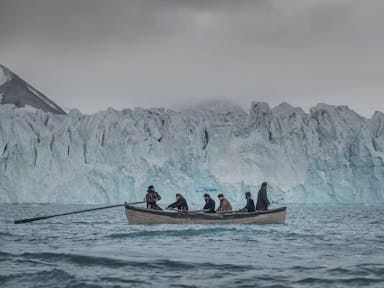 Six men on a small row boat sailing through icy waters past a large glacier in the Arctic