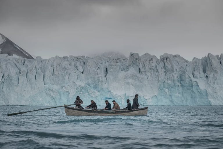 Six men on a small row boat sailing through icy waters past a large glacier in the Arctic