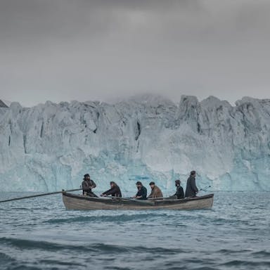 Six men on a small row boat sailing through icy waters past a large glacier in the Arctic
