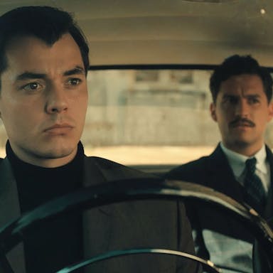 A man in smart clothing sits behind the wheel in a fancy old car, with another man in a suit sat in the back passenger seat