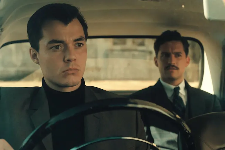 A man in smart clothing sits behind the wheel in a fancy old car, with another man in a suit sat in the back passenger seat