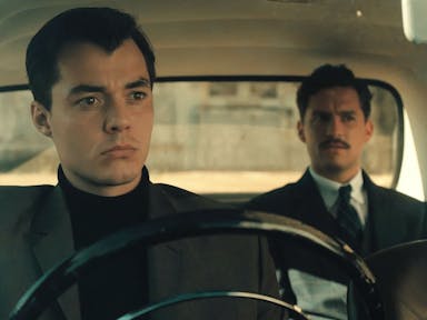 A man in smart clothing sits behind the wheel in a fancy old car, with another man in a suit sat in the back passenger seat
