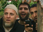 Three men huddled together looking through a gap in the fence looking surprised