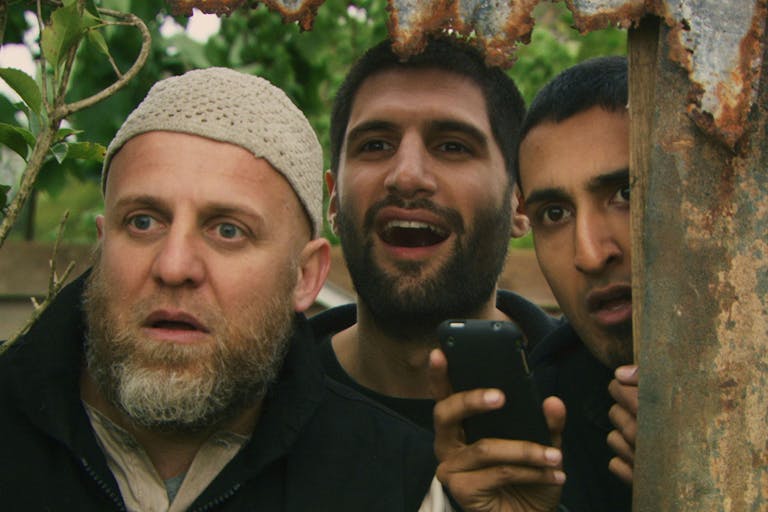 Three men huddled together looking through a gap in the fence looking surprised