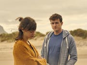 A young woman and young man stand on a windy beach looking thoughtful and melancholic