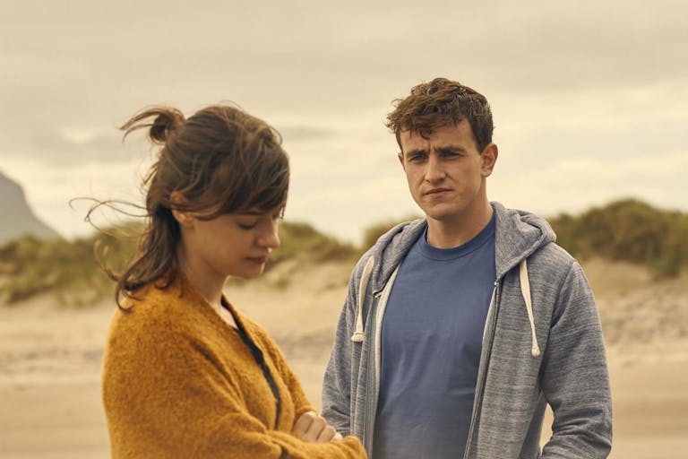 A young woman and young man stand on a windy beach looking thoughtful and melancholic