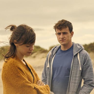 A young woman and young man stand on a windy beach looking thoughtful and melancholic 