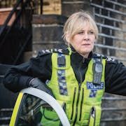 A middle aged white woman in a British police uniform standing in the door of a police car arm resting on door, looking serious into the distance