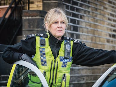 A middle aged white woman in a British police uniform standing in the door of a police car arm resting on door, looking serious into the distance 