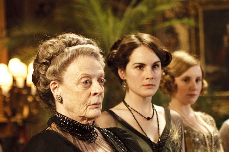 A line of elderly and young white women in regal period clothing in a grand house