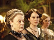A line of elderly and young white women in regal period clothing in a grand house