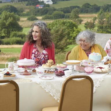 Group of women sit outside in the countryside at a table covered with cakes and teapots