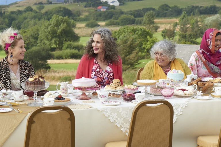 Group of women sit outside in the countryside at a table covered with cakes and teapots