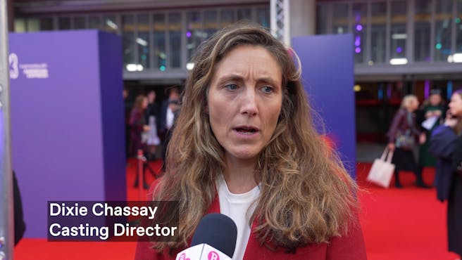 A white woman with long light brown hair and a red blazer talks into a mic on the red carpet