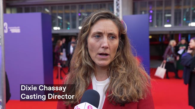 A white woman with long light brown hair and a red blazer talks into a mic on the red carpet