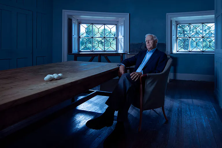 A white older man sitting in a cool blue room by a long wooden table