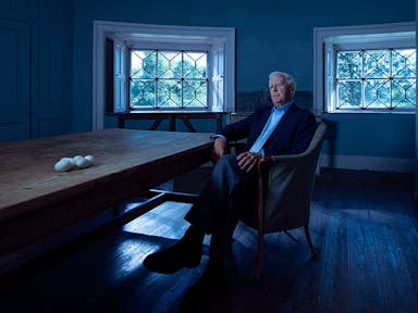 A white older man sitting in a cool blue room by a long wooden table