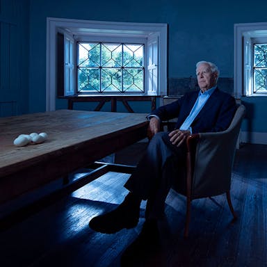A white older man sitting in a cool blue room by a long wooden table