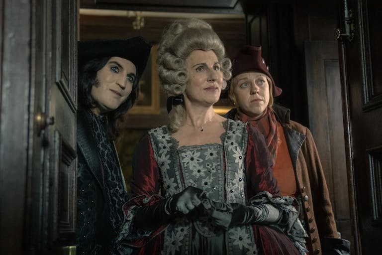 A white man with pale skin, and white woman in a pirate style hat and clothing, and a white women in regal clothing and wig stand in a doorway