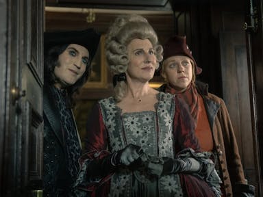 A white man with pale skin, and white woman in a pirate style hat and clothing, and a white women in regal clothing and wig stand in a doorway
