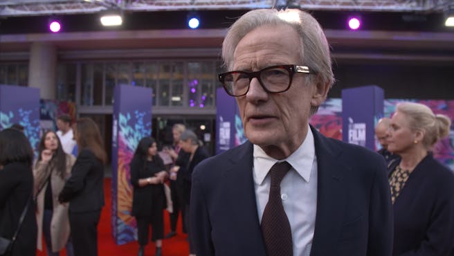 Man with thick-rimmed black glasses wearing a suit on a red carpet.