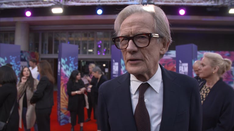 Man with thick-rimmed black glasses wearing a suit on a red carpet. 