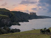 An epic shot of a green coastal hills and cold waters with a grand castle in the distance, a carriage and two horse ride along a path