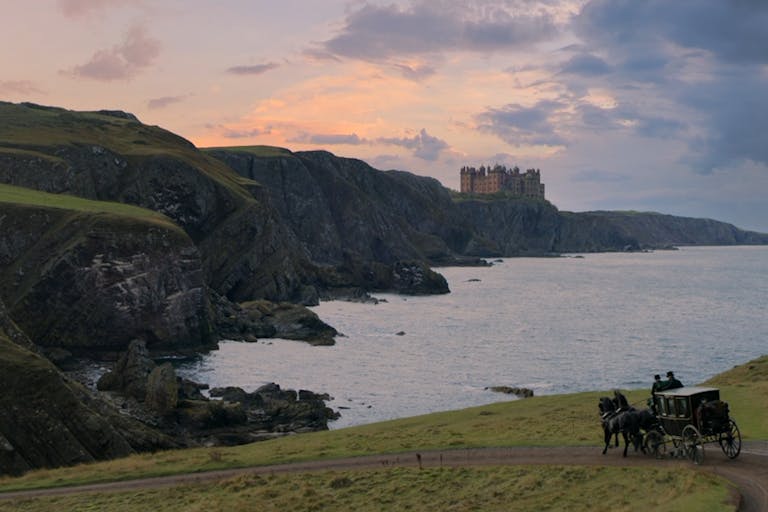 An epic shot of a green coastal hills and cold waters with a grand castle in the distance, a carriage and two horse ride along a path