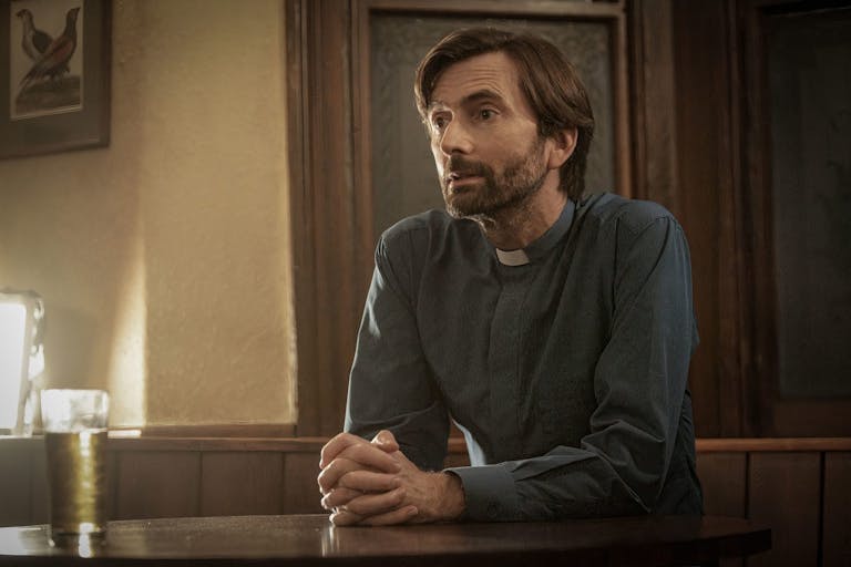 A slim man with dark floppy hair and a trimmed beard wearing a vicar's collar, hands clasped, sitting down with a drink