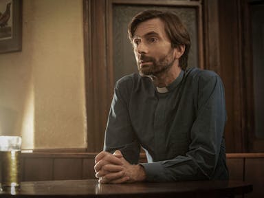 A slim man with dark floppy hair and a trimmed beard wearing a vicar's collar, hands clasped, sitting down with a drink