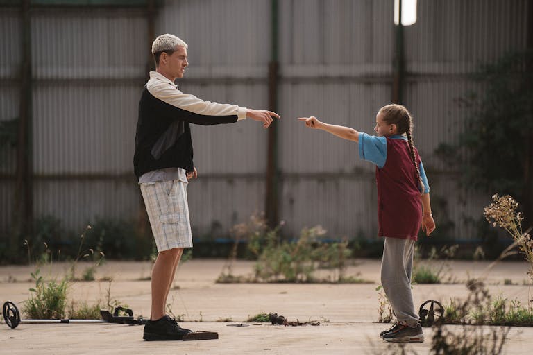 A tall man with bleached hair stands facing a young girl, both pointing at each other in a dance