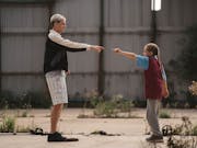 A tall man with bleached hair stands facing a young girl, both pointing at each other in a dance