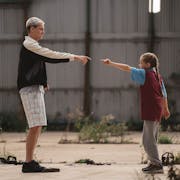 A tall man with bleached hair stands facing a young girl, both pointing at each other in a dance
