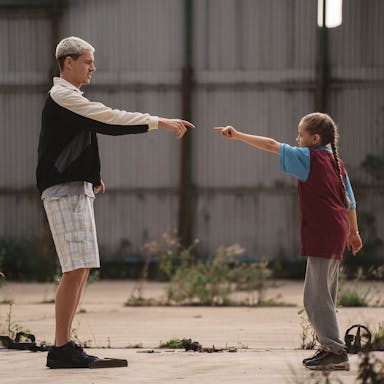 A tall man with bleached hair stands facing a young girl, both pointing at each other in a dance