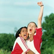 Two young women in a triumphant embrace, arm in the air, both wearing white and red football kit