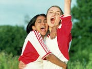 Two young women in a triumphant embrace, arm in the air, both wearing white and red football kit