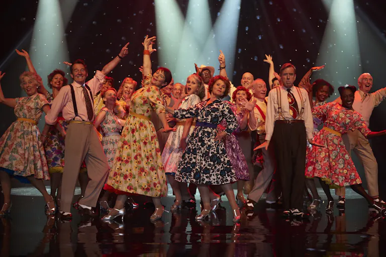 A line of middle aged and older people in swinging dresses and braces on stage dancing
