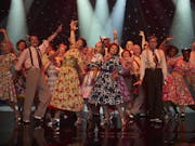 A line of middle aged and older people in swinging dresses and braces on stage dancing