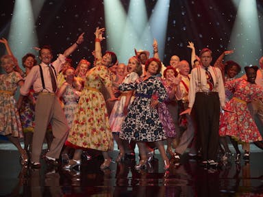 A line of middle aged and older people in swinging dresses and braces on stage dancing 