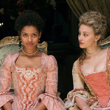 A young mixed-race Black woman and a young white woman dressed in grand period dresses sitting on ornate chairs