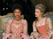 A young mixed-race Black woman and a young white woman dressed in grand period dresses sitting on ornate chairs