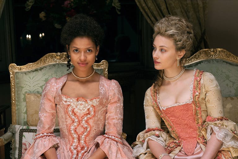 A young mixed-race Black woman and a young white woman dressed in grand period dresses sitting on ornate chairs