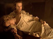 Blonde woman in an old-fashioned white nightgown lies across a man