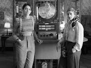 Black and white image of two women standing either side of a slim time machine with lots of dials that they have built