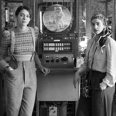 Black and white image of two women standing either side of a slim time machine with lots of dials that they have built 