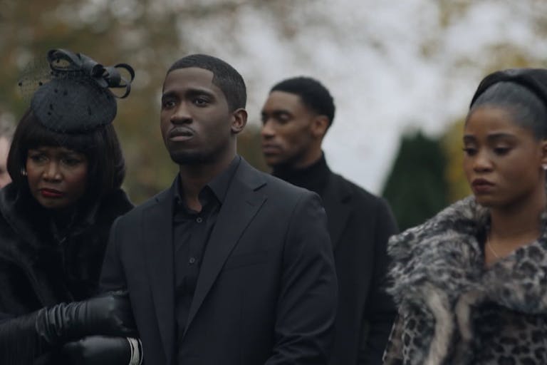 Young man surrounded by a young woman and older woman, dressed in glamourous black outfits with fur, stand outside looking sad. They look like they are at a funeral.